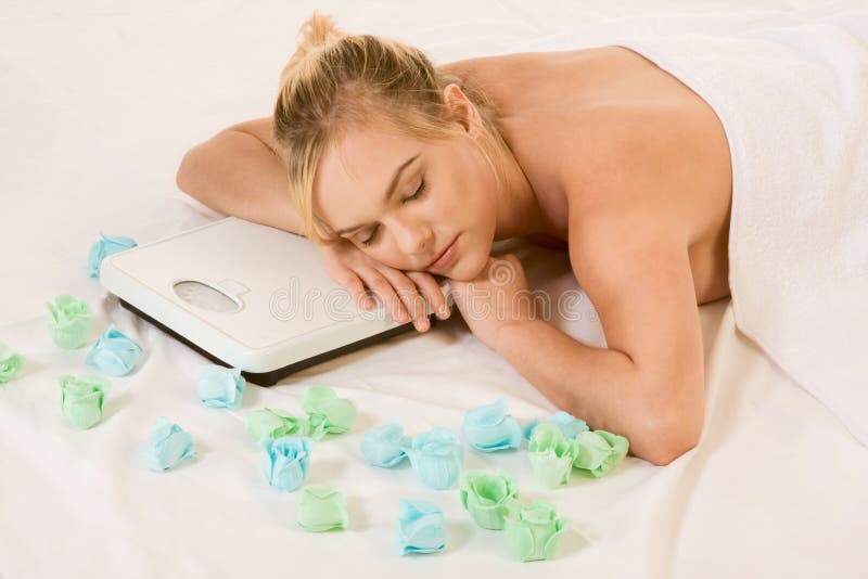 Blonde woman in spa dreaming on weight scales. Blond naked woman lying down awaiting spa treatment, surrounded by aromatherapy items and with her head resting on stock photo