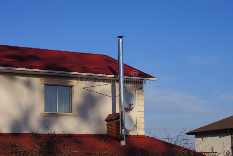 Gray attic of a private house with a window and a metal chimney pipe on a red tile roof against a blue sky stock photos