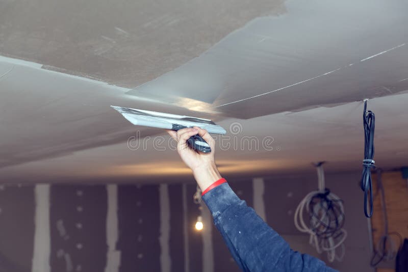 Worker is working with palette-knife for flattening the ceiling from wooden platform in room of apartment is inder stock photos
